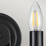 A close-up view of a candle-shaped LED light bulb highlights its visible filament, affixed to a black sconce. Its minimalist design perfectly complements the Capitol Hill 2 Light Wall Light - Black, creating ambient lighting against a neutral backdrop.