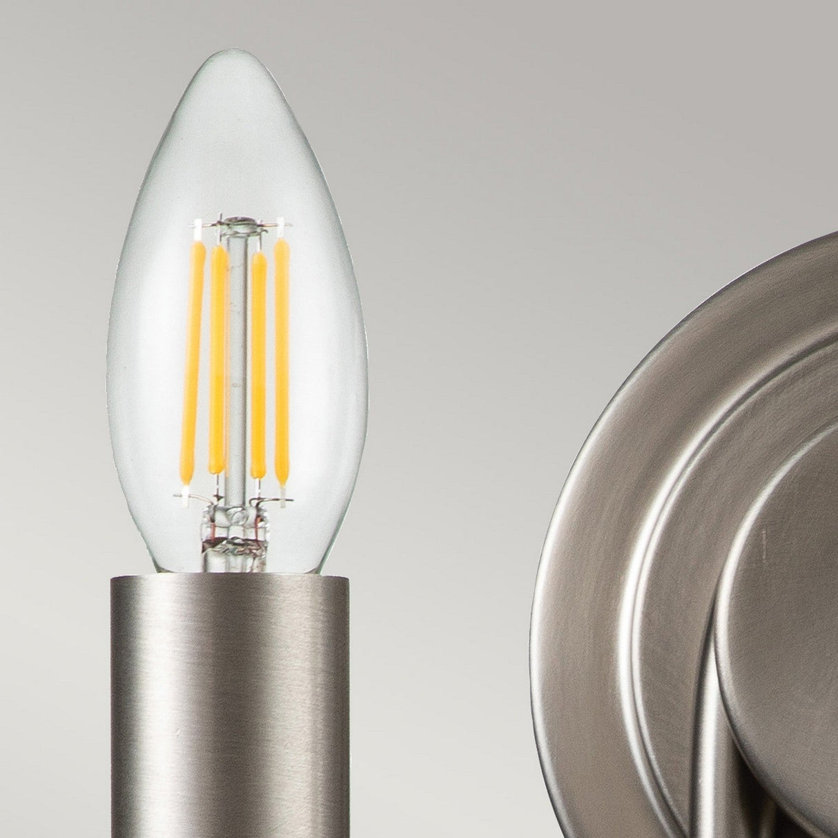 Close-up of a lit LED filament bulb in a candle shape, set in a Capitol Hill 2 Light Wall Light with a brushed nickel holder against a light gray background. The bulb emits a warm glow, partially surrounded by the circular metal fixture.