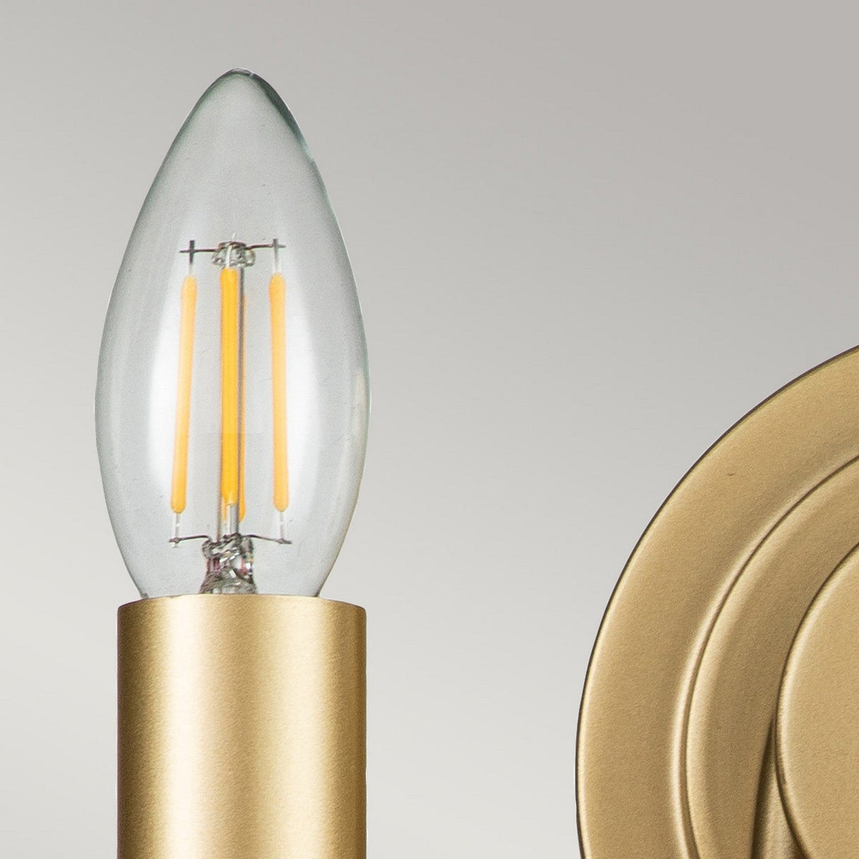 The Capitol Hill 2 Light Wall Light, crafted in natural brass and shaped like a candle with visible filaments, is mounted in a gold-colored metal fixture against a light gray background. This elegant piece of home lighting accentuates the intricate details and texture of both the bulb and its sophisticated fixture.