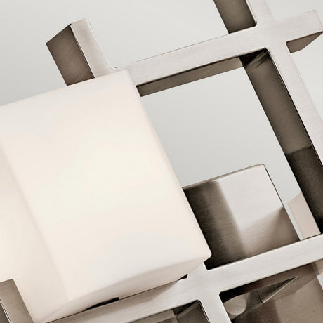 A close-up view of the City Lights 2 Light Wall Light - Silver showcases its modern abstract design, featuring interlocking metal bars with a brushed finish. The smooth, white rectangular block with integrated LED technology stands in contrast to the metallic surfaces. Its contemporary style is enhanced by the plain, light grey background.