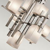 A close-up of the City Lights 7 Light Chandelier - Silver showcases its modern design with an intricate geometric pattern. The chandelier features multiple frosted square glass shades infused with LED lights, artfully arranged on metallic intersecting rods. This arrangement enhances its contemporary and artistic appearance against a neutral background.