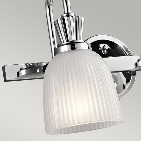 Featuring the Cora 2 Light Wall Light, this modern wall sconce is ideal for bathroom lighting. It boasts a polished chrome fixture and a ribbed frosted glass shade. Designed to be LED-compatible, the lamp is attached to a circular base and emits a soft, warm glow when illuminated.