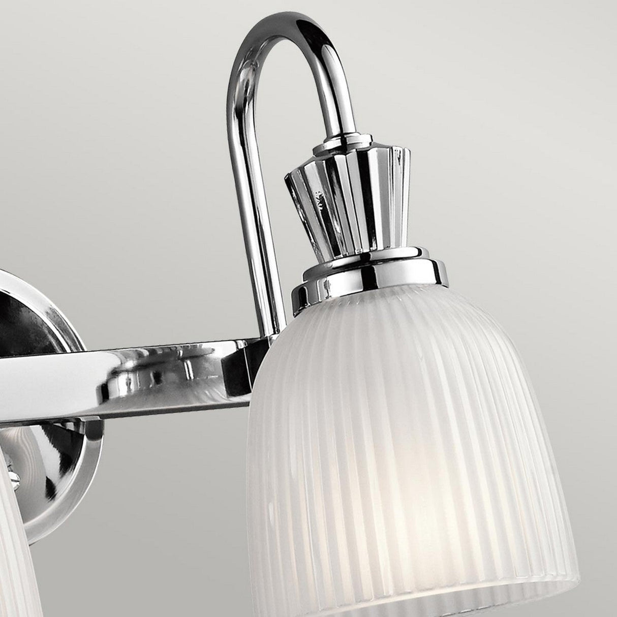 A close-up of the Cora 2 Light Wall Light in polished chrome features a modern wall-mounted fixture with a curved arm and a ribbed frosted glass shade, compatible with LED bulbs, set against a neutral gray background—ideal for bathroom lighting.