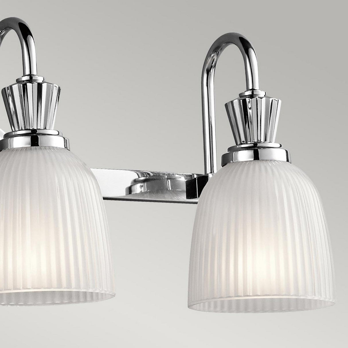 Close-up of a modern three-light vanity fixture with polished chrome accents. This IP44-rated bathroom wall light features ribbed frosted glass shades, each emitting a soft, warm glow against a neutral grey background.