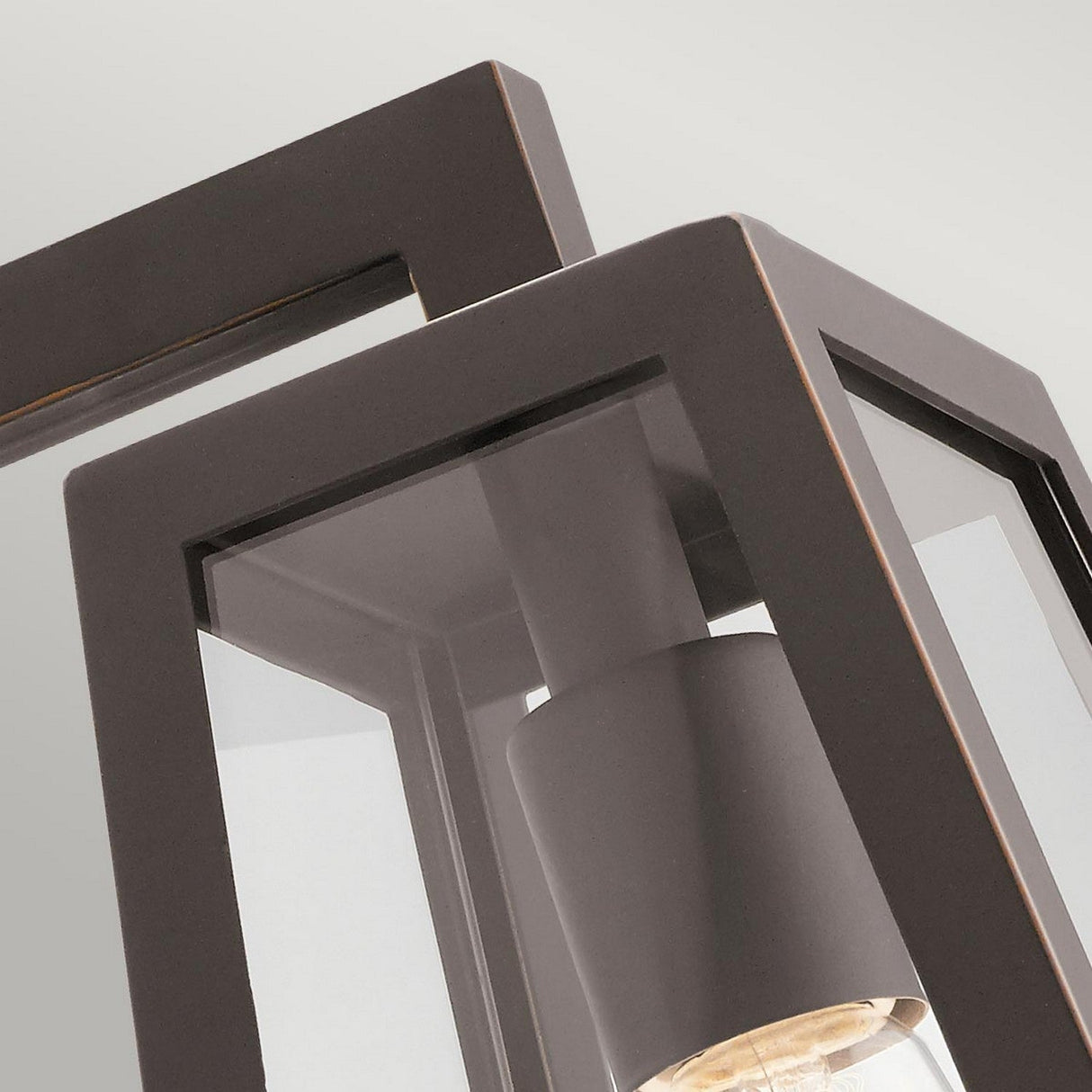 This close-up of the Delison Outdoor 1 Light Wall Lantern in Rubbed Bronze highlights its modern, geometric design with a sturdy bronze frame and clear glass panels, beautifully showcasing the bulb inside. Its angular style brings a contemporary flair to any rustic lodge exterior, offering an elegant contrast against a neutral backdrop while ensuring durability with its IP44 rating.