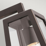 This close-up of the Delison Outdoor 1 Light Wall Lantern in Rubbed Bronze highlights its modern, geometric design with a sturdy bronze frame and clear glass panels, beautifully showcasing the bulb inside. Its angular style brings a contemporary flair to any rustic lodge exterior, offering an elegant contrast against a neutral backdrop while ensuring durability with its IP44 rating.