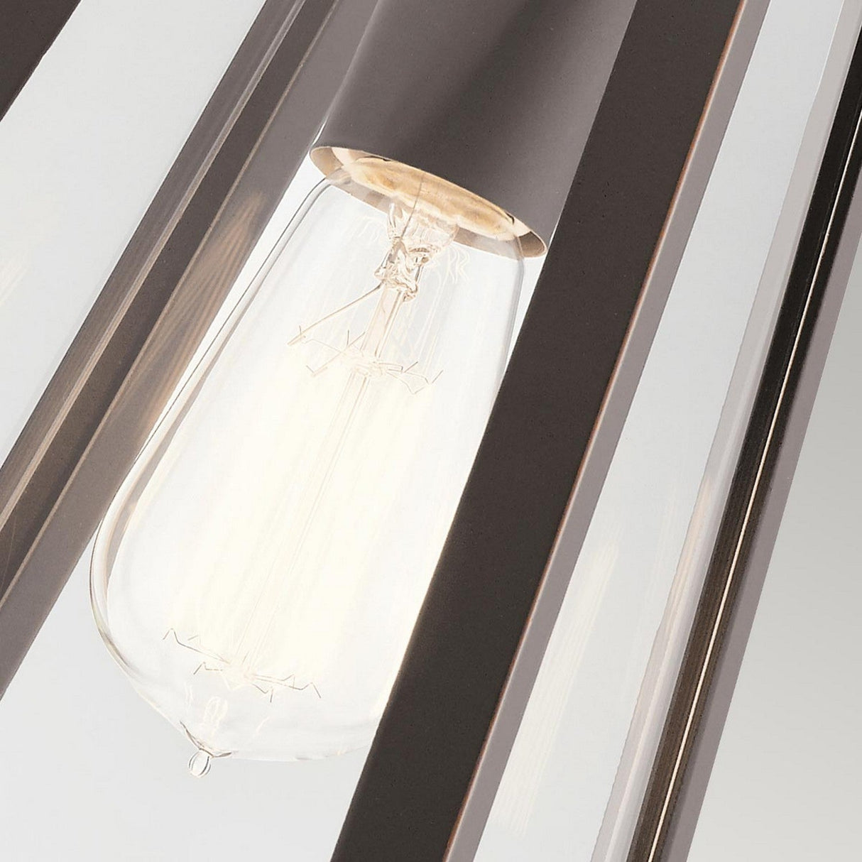 Close-up of the illuminated Delison Outdoor 1 Light Wall Lantern in Rubbed Bronze, featuring a vintage-style bulb set in a sleek metal frame. This IP44-rated outdoor wall light showcases its glowing filament against a soft rustic lodge exterior.