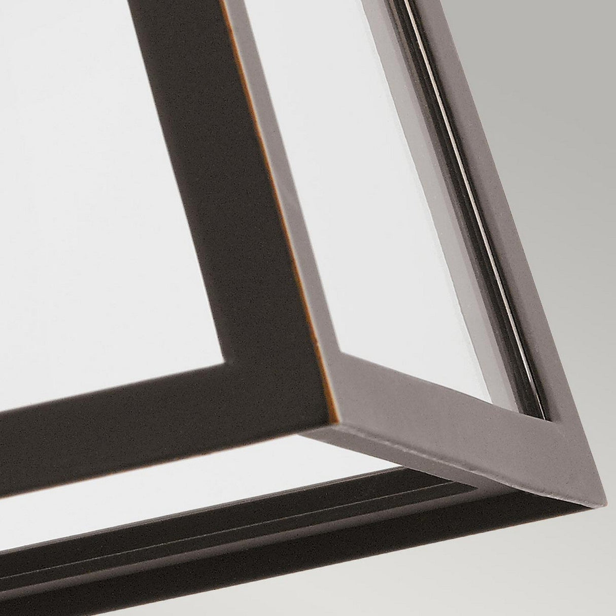 A close-up view of the Delison Outdoor 1 Light Wall Lantern in Rubbed Bronze, illustrating its geometric metal frame with sharp edges and a glass panel. The IP44-rated fixture is captured from below, highlighting its clean lines and modern design against a neutral backdrop.
