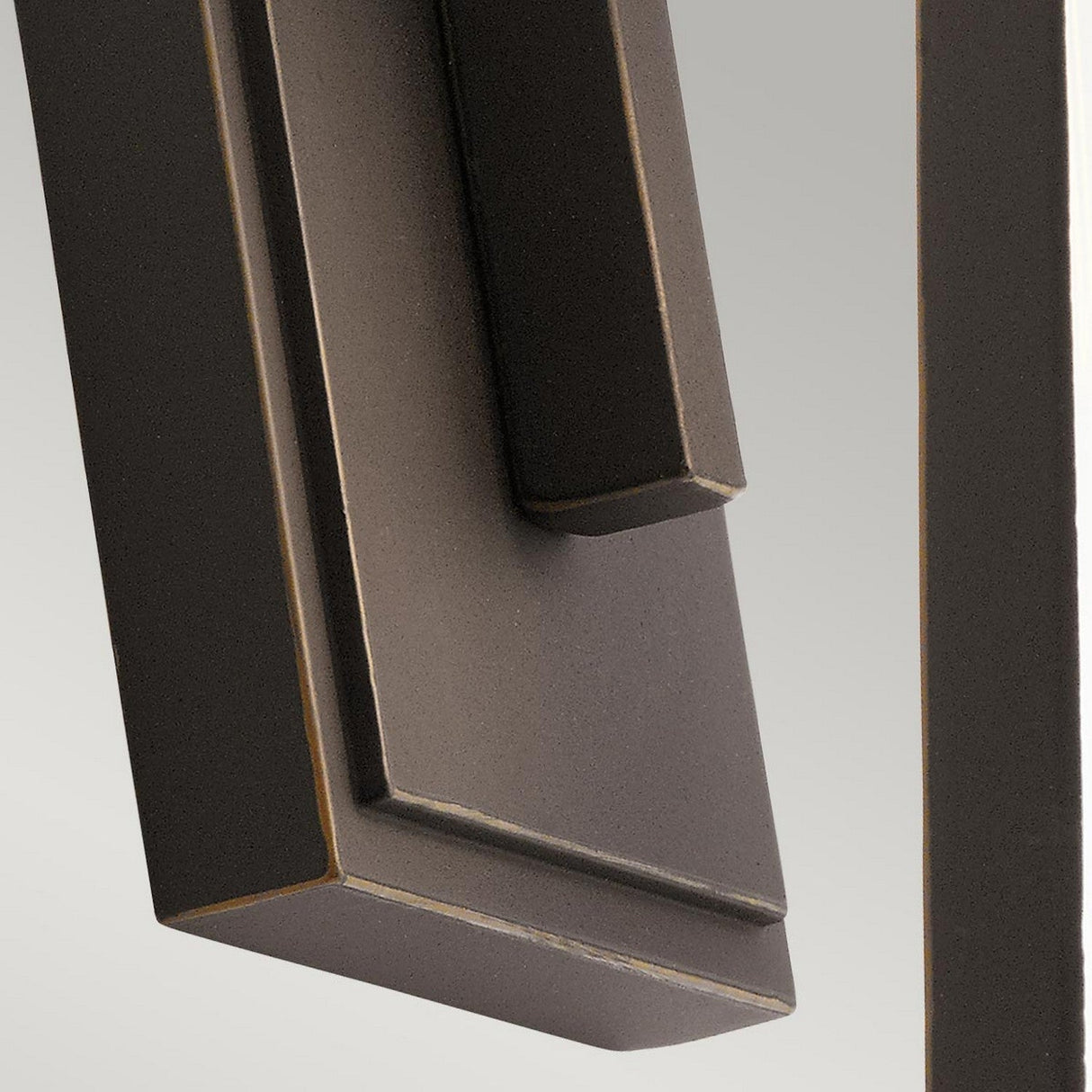 A close-up of a modern, angular metal door handle with a minimalist design, set against a light gray background. The handle, finished in Rubbed Bronze, features clean lines and a matte texture that complements the adjacent Delison Outdoor 1 Light Wall Lantern - Rubbed Bronze (Small), which is IP44-rated for outdoor use.