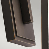 A close-up of a modern, angular metal door handle with a minimalist design, set against a light gray background. The handle, finished in Rubbed Bronze, features clean lines and a matte texture that complements the adjacent Delison Outdoor 1 Light Wall Lantern - Rubbed Bronze (Small), which is IP44-rated for outdoor use.