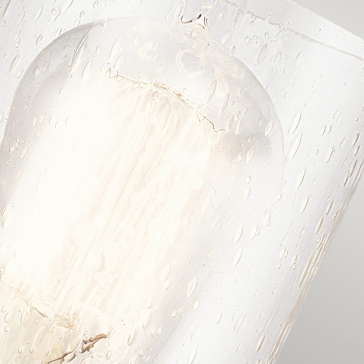 A detailed view of the Deryn 1 Light Wall Light in Antique Grey reveals a lit bulb inside a glass enclosure adorned with water droplets, producing a diffused and soft glow. This image captures rustic elegance through its tranquil and calming ambiance, accentuated by how the light interacts with the clear seeded glass.