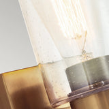 Close-up of the Deryn 1 Light Wall Light in a modern design, showcasing a clear, seeded glass shade accented with subtle bubbles. A glowing filament bulb inside emits a warm ambience, and the sleek natural brass finish harmonizes beautifully with both contemporary and rustic decor styles.