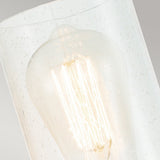 The Deryn 1 Light Wall Light in Natural Brass features a close-up view of a glowing filament bulb inside its clear seeded glass fixture, adorned with small air bubbles. This light casts a warm and inviting glow that beautifully highlights the intricate details of the bulb's design, making it an ideal choice for rustic decor.