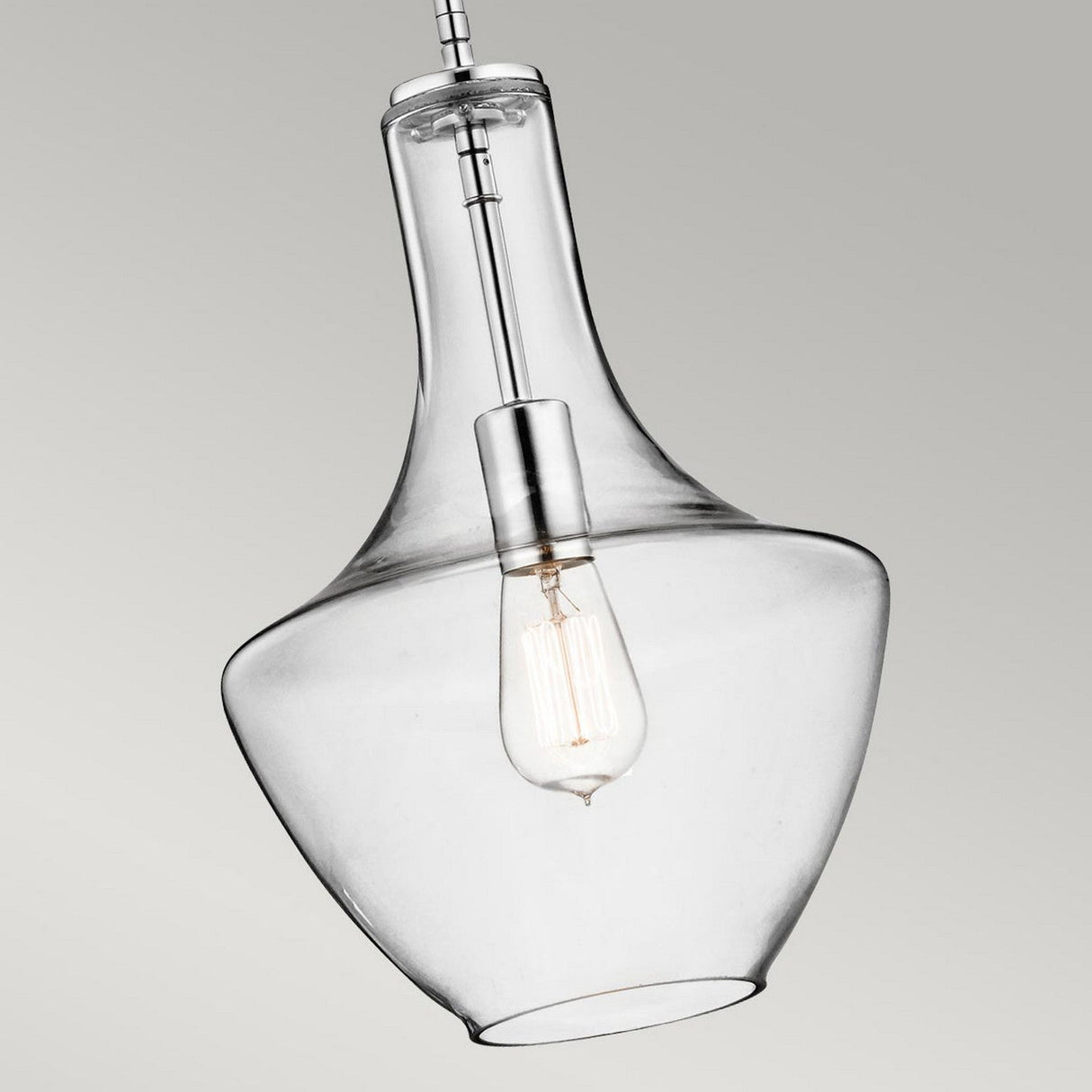 A close-up view of the Everly 1 Light Small Pendant in chrome showcases its clear glass shade with a visible bulb inside. The pendant features a sleek, curved design and is suspended from a chrome-finished metal rod, embodying vintage-inspired decor against a neutral backdrop.