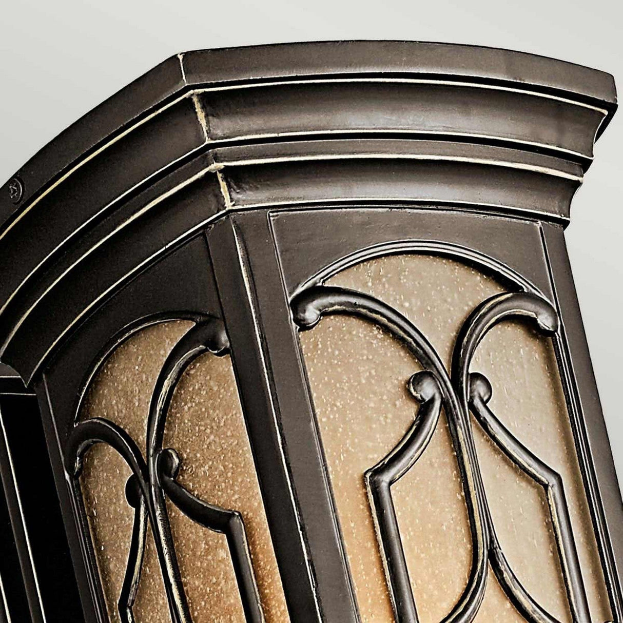A close-up of the Franceasi Outdoor 1 Light Medium Wall Lantern in Olde Bronze showcases its intricate wrought iron design with curved panels and warm amber-tinted glass. Set against a light, neutral background, the elegant metalwork is beautifully emphasized.