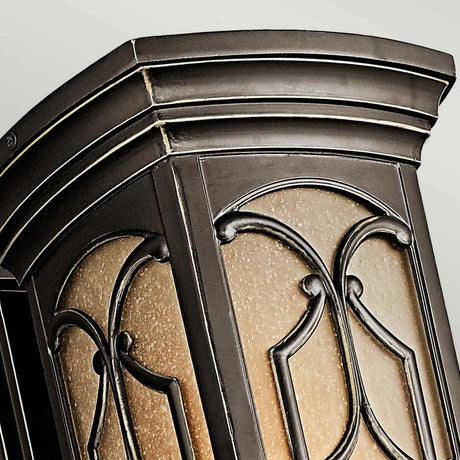 A close-up of the Franceasi Outdoor 1 Light Medium Wall Lantern in Olde Bronze showcases its intricate wrought iron design with curved panels and warm amber-tinted glass. Set against a light, neutral background, the elegant metalwork is beautifully emphasized.