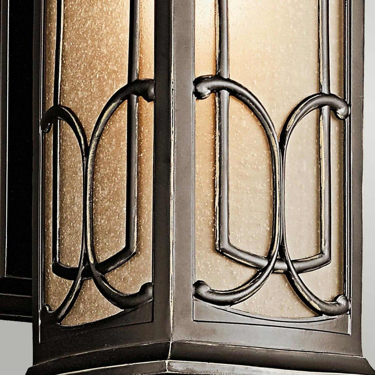 A detailed view of the Franceasi Outdoor 1 Light Medium Wall Lantern reveals its amber-tinted glass panels and elaborate black metal scrollwork. The gentle illumination from within casts a warm glow that accentuates the textured glass and graceful design, making it an ideal choice for adding charm with its olde bronze finish.