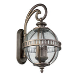 Introducing the Halleron Outdoor 3 Light Wall Lantern, a fixture that exudes Victorian elegance with its vintage-style design. It features a clear, spherical glass cover enhanced by a decorative burnished bronze finish. This striking lighting piece is attached to a curved metallic arm and mounted on a rectangular backplate.