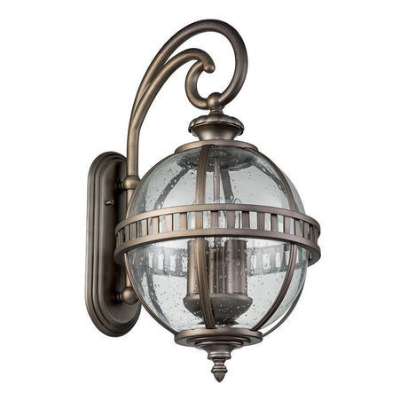 Introducing the Halleron Outdoor 3 Light Wall Lantern, a fixture that exudes Victorian elegance with its vintage-style design. It features a clear, spherical glass cover enhanced by a decorative burnished bronze finish. This striking lighting piece is attached to a curved metallic arm and mounted on a rectangular backplate.