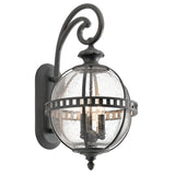 Introducing the Halleron Outdoor 3 Light Wall Lantern in Grey, showcasing Victorian sophistication with its clear seeded glass globe and black metal accents in an ornate, curved pattern. This vintage design radiates classic charm and is elegantly mounted on a rectangular backplate.