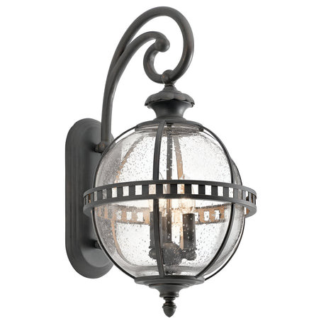 Introducing the Halleron Outdoor 3 Light Wall Lantern in Grey, showcasing Victorian sophistication with its clear seeded glass globe and black metal accents in an ornate, curved pattern. This vintage design radiates classic charm and is elegantly mounted on a rectangular backplate.