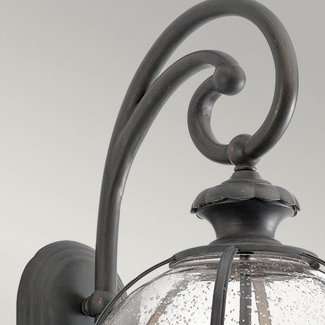 Close-up of the Halleron Outdoor 3 Light Wall Lantern, showcasing its vintage-style charm with a dark metal finish. This lantern features a decorative scroll arm and a clear glass shade with a bubbled texture, set against a simple light grey background.