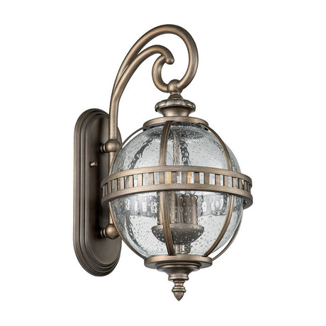 Illuminate your outdoor space with the timeless elegance of the Halleron Outdoor 2 Light Wall Lantern in a burnished bronze finish. This gorgeous fixture showcases a spherical, clear seeded glass shade and intricate scrollwork, all beautifully supported by a decorative curved arm and backplate.