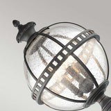 A close-up of the Halleron Outdoor 3 Light Pedestal Lantern showcases its Victorian-inspired, spherical design. The fixture features a black metal frame with textured glass panels that reveal a visible bulb inside. With an IP44 rating, this lantern contrasts beautifully against the plain gray background.