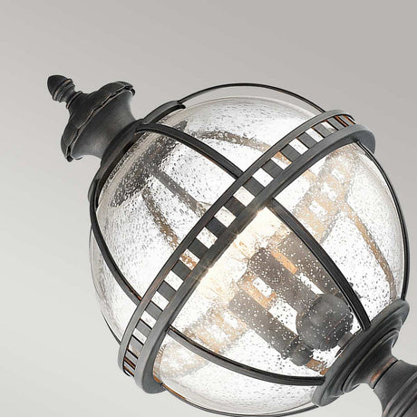 A close-up of the Halleron Outdoor 3 Light Pedestal Lantern showcases its Victorian-inspired, spherical design. The fixture features a black metal frame with textured glass panels that reveal a visible bulb inside. With an IP44 rating, this lantern contrasts beautifully against the plain gray background.