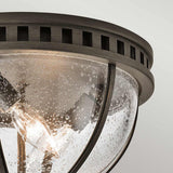 A close-up of the Halleron Outdoor 3 Light Ceiling Flush - Bronze reveals its metal frame paired with a clear, bubbled glass dome that enhances its Victorian design. The bronze finish complements this aesthetic, while inside, three glowing bulbs cast a warm glow from the plain, light-colored ceiling.