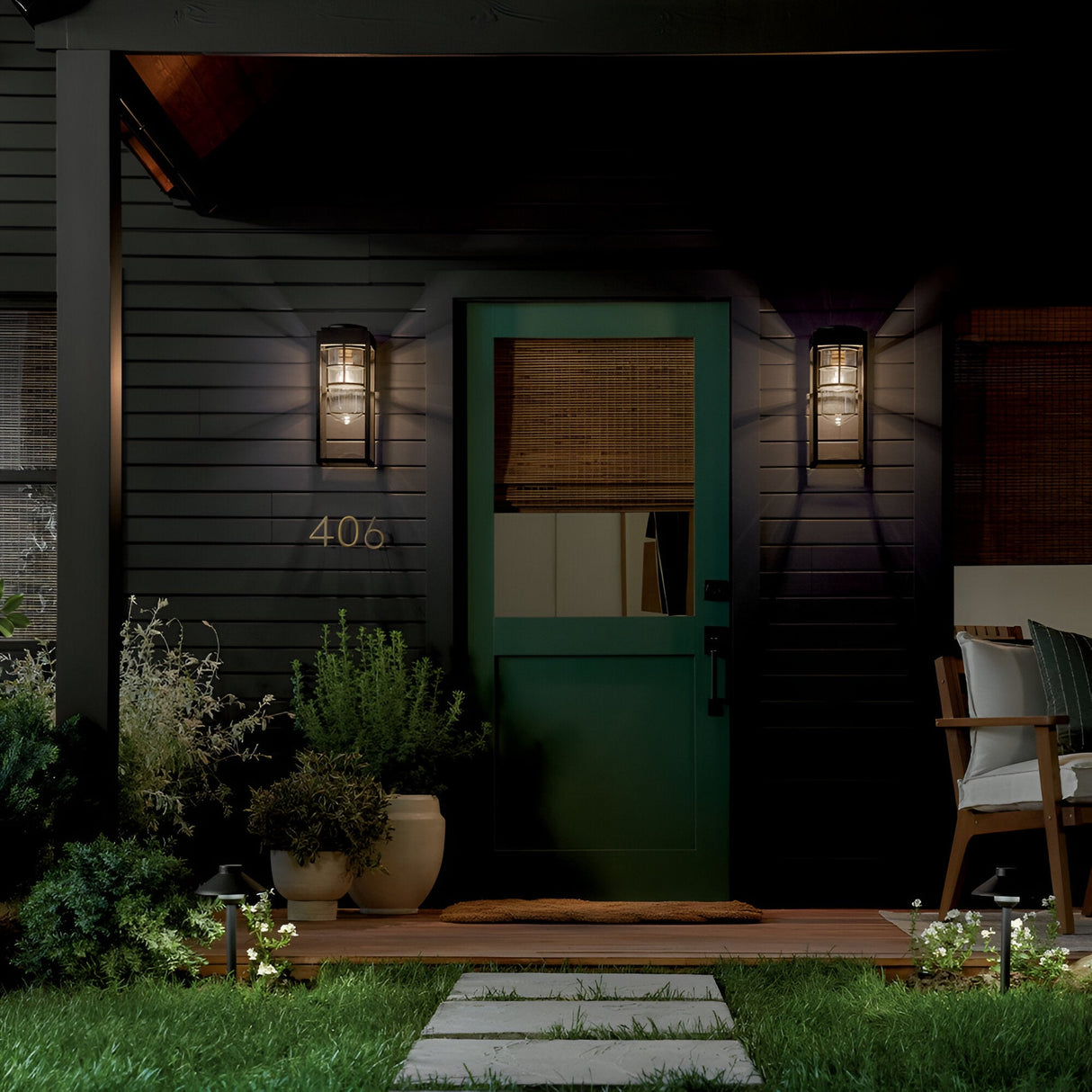 A green front door with glass panels is framed by Hone Coastal 1 Light Medium Outdoor Wall Lanterns in black on a dark house. Potted plants, a doormat, house number 406, and a stone path across the grass complete the entryway.