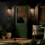 A green door with a window and bamboo shade is flanked by Hone Coastal 1 Light Medium Outdoor Wall Lanterns in black. A welcome mat, potted plants, and a cushioned chair with pillows complete the cozy porch setting for evening relaxation.
