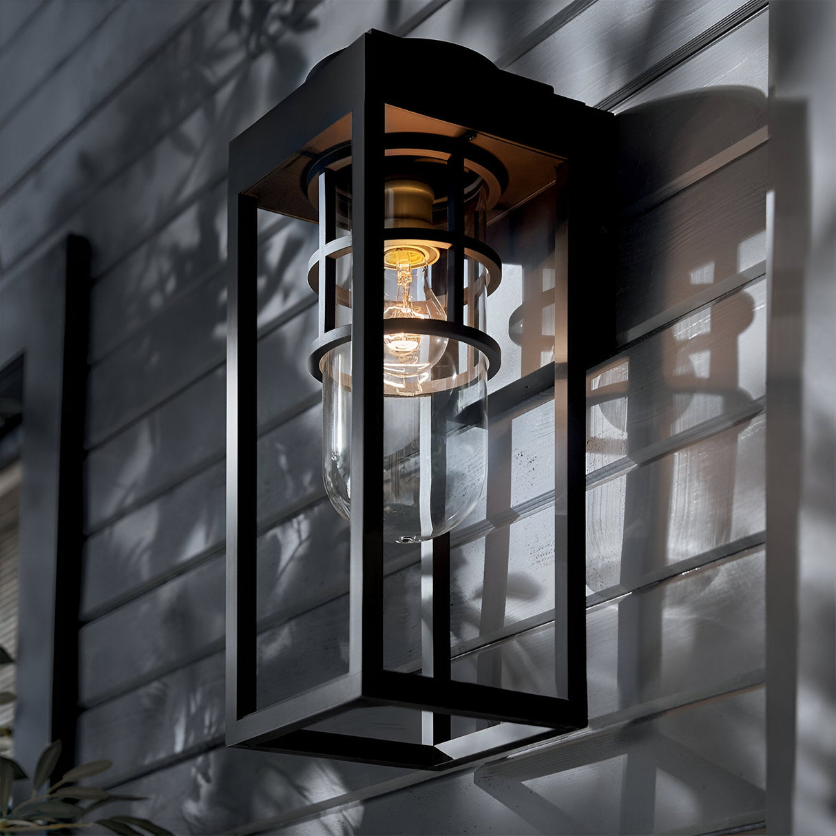 The Hone Coastal 1 Light Medium Outdoor Wall Lantern in black is installed on gray horizontal siding, offering weather-resistant illumination. Glass enclosure casts striking shadows from nearby foliage along the brightened wall.