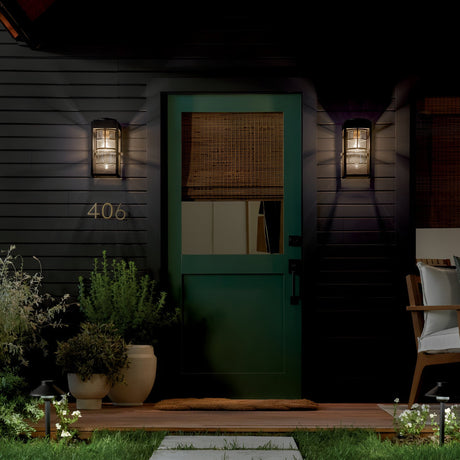 A green front door with a woven shade is lit at night by the Hone Coastal 1 Light Small Outdoor Wall Lantern in black. House number 406 and a wooden chair sit on the porch, surrounded by plants and grass.