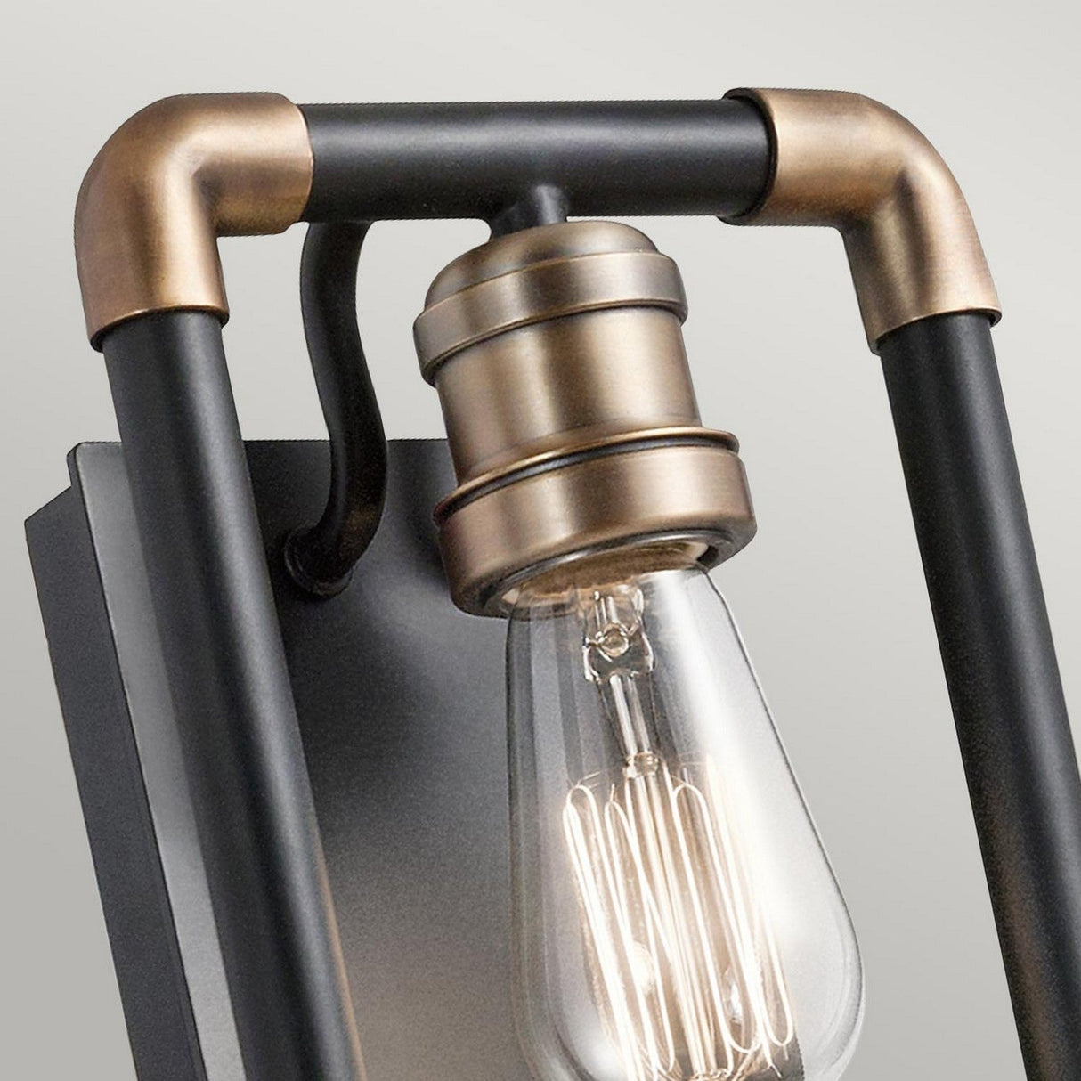 Close-up of the Imahn 1 Light Wall Light in black and natural brass featuring an Edison-style bulb. The fixture's minimalist design, with dark metal rods and brass accents, enhances the industrial look against a neutral background.