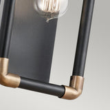 A close-up of the Imahn 1 Light Wall Light showcases its industrial style with black metal rods and natural brass connectors. An Edison-style bulb emits a warm glow, enhancing the minimalist design against a neutral, light gray background.