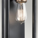 Close-up of an Edison-style bulb with visible filament, set in an Imahn 1 Light Wall Light featuring a sleek black and natural brass fixture against a neutral background. The design emphasizes the bulb as the focal point, showcasing its minimalist and industrial style.