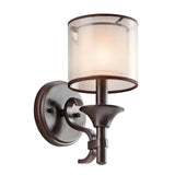 The Lacey 1 Light Wall Light in Bronze is a wall-mounted fixture with a rustic finish, featuring a circular base and an elegantly curved arm. It includes a cylindrical frosted glass shade that gently diffuses light, and it offers an optional double-shade design in organza fabric to enhance its elegance.