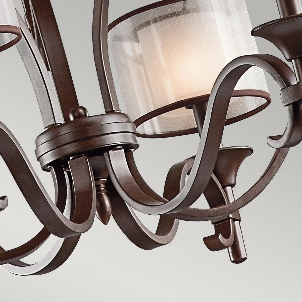 Close-up of the Lacey 9 Light Chandelier - Bronze, showcasing its ornate double-shade design with elegantly curved metal arms and frosted glass shades, each illuminated by a bulb, set against a plain gray background.