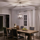 A dining room with a wooden table and six chairs features the Larkin 3 Light Medium Chandelier in Brushed Nickel hanging above. The open cage design of the chandelier adds elegance to the decor, with woven baskets, a vase, and a bowl of green apples adorning the table. White curtains cover the windows.