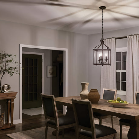 The cozy dining room showcases a wooden table surrounded by six chairs, all illuminated by the Larkin 3 Light Medium Chandelier in Olde Bronze. The chandelier's open cage design brings a touch of elegance to the space, enhanced by decorative vases and a bowl of green apples. Soft lighting highlights the wooden floors, framed gently by delicate curtains.