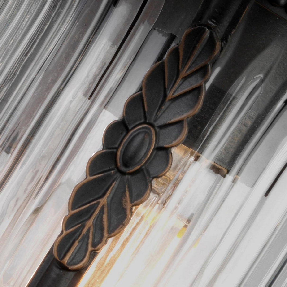 A close-up of the Luverne Outdoor 1 Light Small Wall Lantern in rubbed bronze, featuring a leaf and oval design. The fixture is adorned with clear, vertically ribbed glass panels that house a warmly glowing light bulb.