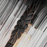 A close-up of the Luverne Outdoor 1 Light Small Wall Lantern in rubbed bronze, featuring a leaf and oval design. The fixture is adorned with clear, vertically ribbed glass panels that house a warmly glowing light bulb.