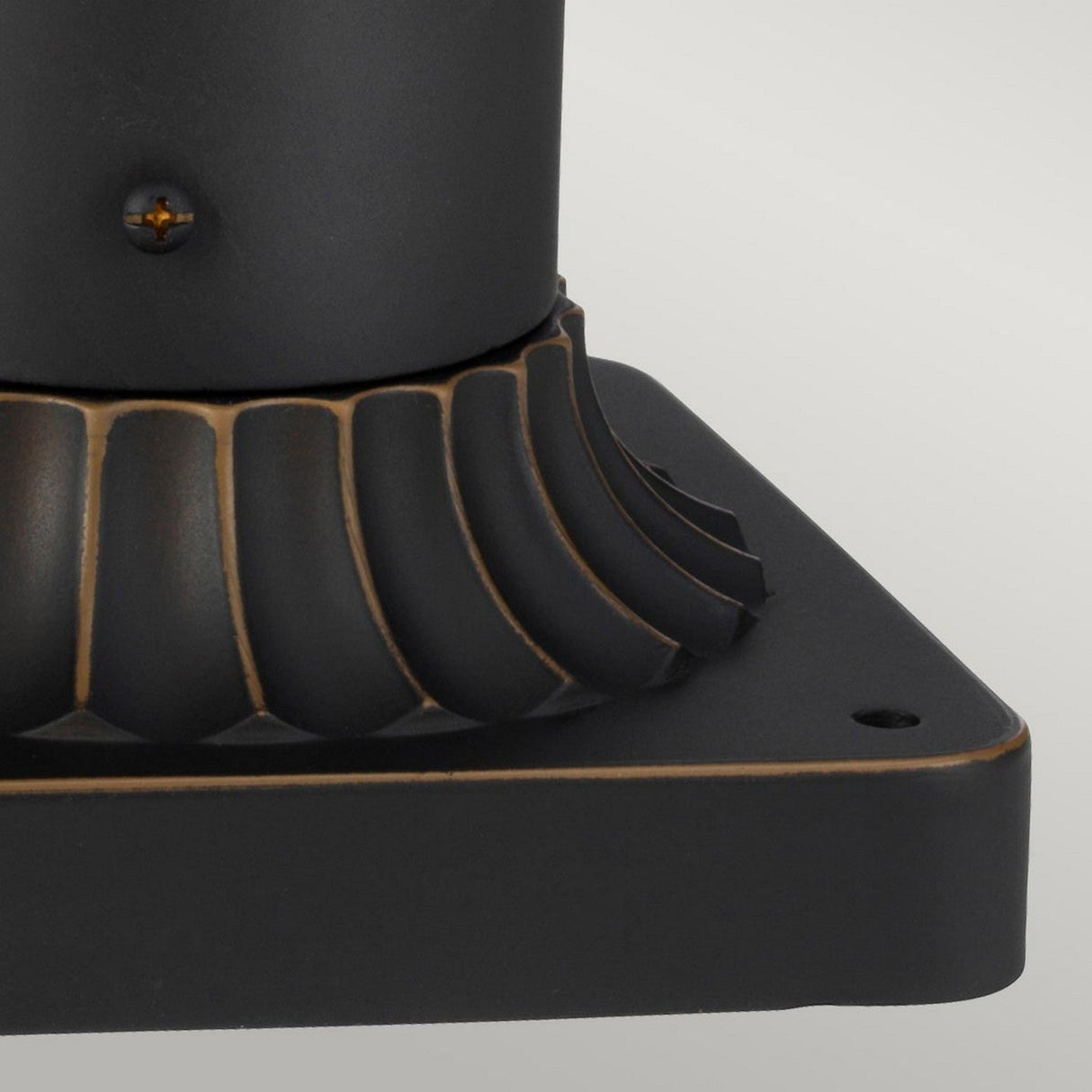 The Luverne Outdoor 1 Light Medium Pedestal in rubbed bronze features a close-up of its black metal base adorned with a decorative ridged design. Its rectangular base, boasting a smooth finish, supports a central cylindrical structure. Subtle highlights accentuate the curves and edges, making it an ideal choice for an outdoor pedestal light.