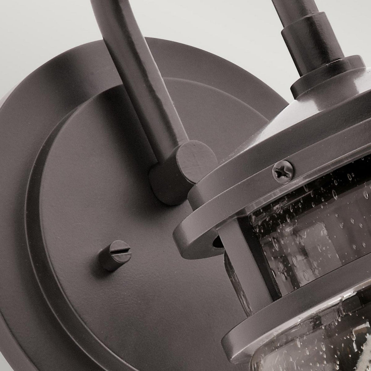 The Lyndon Outdoor 1 Light Medium Wall Lantern, featuring a sleek bronze finish, showcases a round backplate and a seedy glass shade that mimics water droplet details, securely held by screws. A robust metal arm elegantly holds the lantern in place.