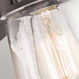 A close-up of the Lyndon Outdoor 1 Light Small Wall Lantern reveals a vintage-style light bulb nestled inside a clear seedy glass fixture speckled with small bubbles. The bulb casts a warm glow, while the lantern is mounted on an elegant metallic base featuring a bronze finish.