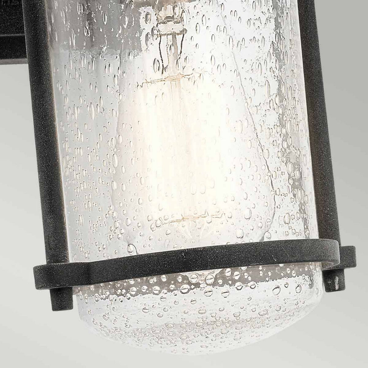 A close-up of the Riverwood Outdoor 1 Light Medium Wall Lantern - Weathered Zinc showcases rustic elegance with its black metal frame and clear, textured glass housing. The bubble-like texture enriches the soft glow against a neutral gradient, while its weathered zinc finish adds timeless charm.