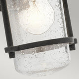 The Riverwood Outdoor 1 Light Wall Lantern, featuring a glowing bulb within its textured clear glass and encased in a weathered zinc metal frame with visible bubbles, brings to mind a rustic seaside atmosphere infused with nautical inspiration and charm.