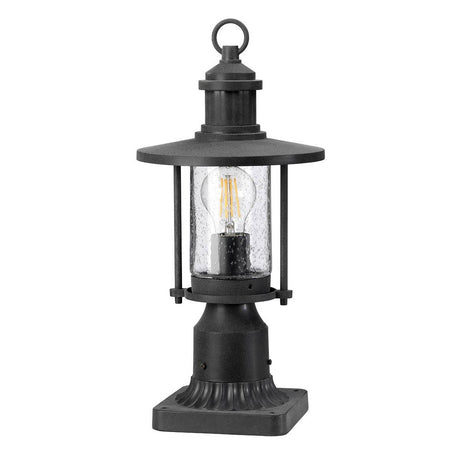 The Riverwood Outdoor 1 Light Pedestal Lantern - Weathered Zinc features a nautical-inspired design with a clear glass case and visible LED bulb. It has a flat, round top and decorative square base, making it ideal for garden or pathway lighting. Its weathered zinc finish adds timeless charm to any outdoor space.