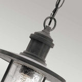 Close-up of the Riverwood Outdoor 1 Light Chain Pendant, showcasing its rustic, weathered zinc finish. The pendant boasts a clear glass enclosure with a textured, wavy surface and is suspended from a chain against a simple, light gray background, capturing the charm of nautical pendant lighting.